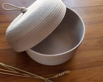 Puede incluir: Un cuenco de cuerda beige con una tapa a juego, parcialmente abierta. El cuenco y la tapa están hechos de cuerda enrollada, creando una superficie texturizada. Un asa de cuerda está unida a la tapa. El cuenco está sobre una superficie de madera, con espigas de trigo secas en primer plano.