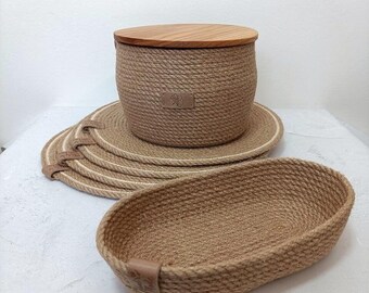 Puede incluir: Un conjunto de artículos de decoración para el hogar de yute tejido. Incluye una cesta redonda con tapa de madera, una pequeña cesta ovalada y varios manteles individuales redondos. Los artículos son beige y marrón, con detalles en cuero.