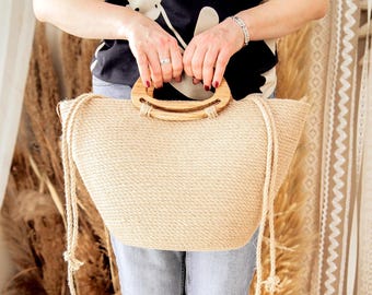 May include: A beige woven tote bag with a wooden handle and rope straps. The bag is held by a person wearing a black and white floral top and blue jeans. The bag has a natural, rustic appearance.