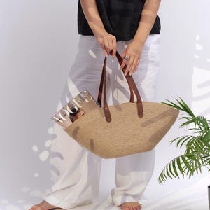 May include: A woven straw tote bag with brown leather handles. The bag is held by a person wearing a black top and white pants. A magazine is visible inside the bag. The bag is a neutral color and has a wide, open design.