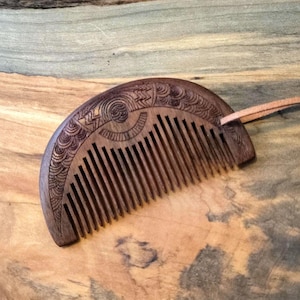 May include: A half-moon shaped wooden comb with a decorative carved design and a leather strap. The comb is made of dark brown wood and has a natural finish.