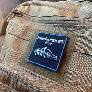 Puede incluir: Parche cuadrado azul marino con el texto "Professional WEE WOO driver" y un coche de policía. El parche tiene un borde azul claro y está adherido a una mochila táctica de color beige.