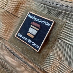 Puede incluir: Un parche cuadrado negro con el texto "Running on Caffeine And Probable Cause" y un gráfico de taza de café. El parche tiene bordados blancos y marrones. Está adherido a un fondo de tela beige.