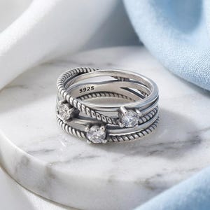 May include: A silver ring with three bands, featuring a braided rope design and three clear, round gemstones. The ring is marked with "S925" and sits on a white marble surface. The background includes white and blue fabric.