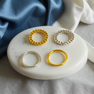 May include: Four rings on a white oval surface. Two are gold, one silver, and one a gold chain link design. The rings are set against blue and white fabric. The rings are suitable for everyday wear.