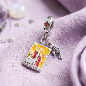 May include: A silver charm featuring a rectangular tarot card pendant labelled THE MAGICIAN with yellow and red enamel, an infinity symbol, and a small pink stone. It includes a separate silver hand charm and a bail decorated with a sun and orange stone.