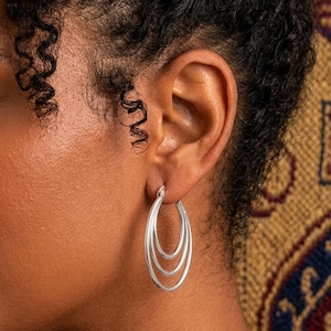 May include: Silver triple hoop earrings. The earrings are oval-shaped and consist of three nested hoops. The earrings are worn on a person's ear, with the skin tone appearing light brown. The background is a patterned fabric.