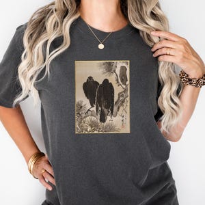 Puede incluir: Camiseta gris oscuro con una impresión enmarcada de dos pájaros negros posados en una rama. La obra de arte es de estilo japonés tradicional, con tonos negros, marrones y beige. La camiseta tiene un corte holgado y mangas cortas.