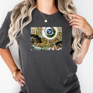 May include: Dark gray t-shirt with a colorful graphic print. The graphic features a circular design with a landscape scene, including trees, water, and figures. The shirt has short sleeves and a crew neck.
