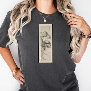 Op de afbeelding: Donkergrijs T-shirt met een ingelijst, verticaal landschapsschilderij. Het kunstwerk toont een bergachtig tafereel met bomen en een rivier, in gedempte tinten. Het shirt heeft een relaxte pasvorm en korte mouwen.