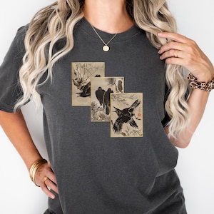 May include: Dark gray t-shirt featuring three framed images of crows in a Japanese art style. The images are in shades of black, brown, and beige. The shirt has short sleeves and a relaxed fit.
