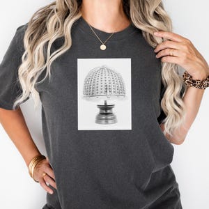 Può includere: T-shirt grigio scuro con un'immagine in bianco e nero di una lampada vintage. La lampada ha un paralume a cupola con piccoli dettagli distanziati uniformemente e una base a piedistallo. La maglietta è uno stile casual.