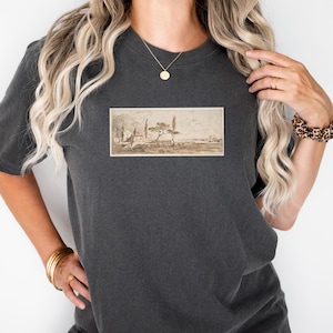 May include: Dark gray t-shirt featuring a sepia-toned landscape print. The artwork depicts trees, buildings, and a distant mountain range. The shirt has short sleeves and a relaxed fit, suitable for casual wear.