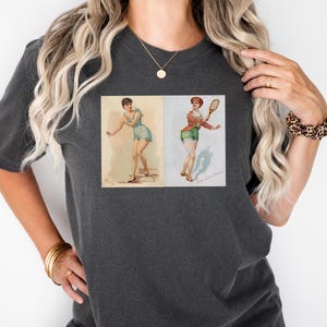 Op de afbeelding: Donkergrijs T-shirt met een vintage illustratie van twee vrouwen die tennis spelen. De illustraties zijn in retrostijl, met een vrouw in een blauwe outfit en de andere in een rood en groen outfit. Het shirt is een comfortabele, casual stijl.