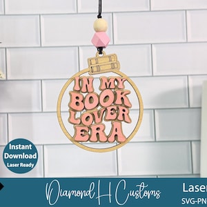 May include: A wooden ornament with the phrase "IN MY BOOK LOVER ERA" in pink lettering. The ornament is circular with a gold border and features a small stack of books at the top. It hangs from a black string with wooden and pink beads.