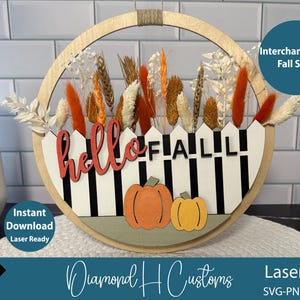 Könnte beinhalten: Runder Holz-Herbst-Dekoschild mit weißem Zaun-Design, den Worten "hello FALL" in Rot und Schwarz und zwei orangefarbenen Kürbissen. Das Schild ist mit Trockenblumen und Weizen verziert. Laserfertig.