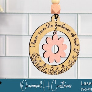 May include: Wooden laser-cut ornament with a floral design and the phrase "I have seen the goodness of God." The ornament features a pink flower in the center and is suspended by a wooden bead and a pink geometric bead. The design includes floral accents.