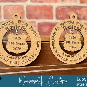 May include: Two wooden Route 66 ornaments. Each ornament features the text "Route 66", the years "1926" and "2026", and the words "100 Years". The ornaments also list the cities and states along the route. The text "2,448 Miles" is also included.