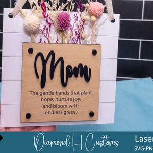 May include: A wooden sign with the word "Mom" in black lettering and a quote about the gentle hands that plant hope, nurture joy, and bloom with endless grace. The sign is decorated with dried flowers.