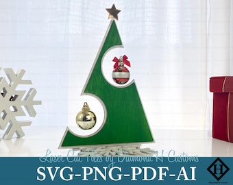 May include: A laser cut wooden Christmas tree with a green painted surface and three ornaments. The tree is decorated with a gold star on top and two round ornaments, one gold and one red with white stripes. The text "Laser Cut Trees by Diamond H Customs" and "SVG-PNG-PDF-AI" are visible at the bottom of the image.