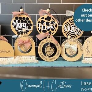 May include: Wooden laser-cut ornaments with the words "Mom" and "Mama" in various designs. Some ornaments have decorative bows and macrame fringe. The image also includes the text "Check out our other designs."