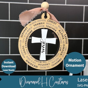 May include: Wooden ornament featuring a white cross with the word "Jesus" in script. The cross is set within a circular frame with religious terms. A pink ribbon and wooden bead are attached for hanging. The words "Motion Ornament" are also visible.