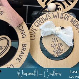 May include: Three wooden ornaments with laser cut designs. The first ornament says "Love Grows Where Mom Goes!" with a white ribbon and a floral design. The second ornament says "Song Brave" with a heart design. The third ornament says "He is Mom" with a heart design.