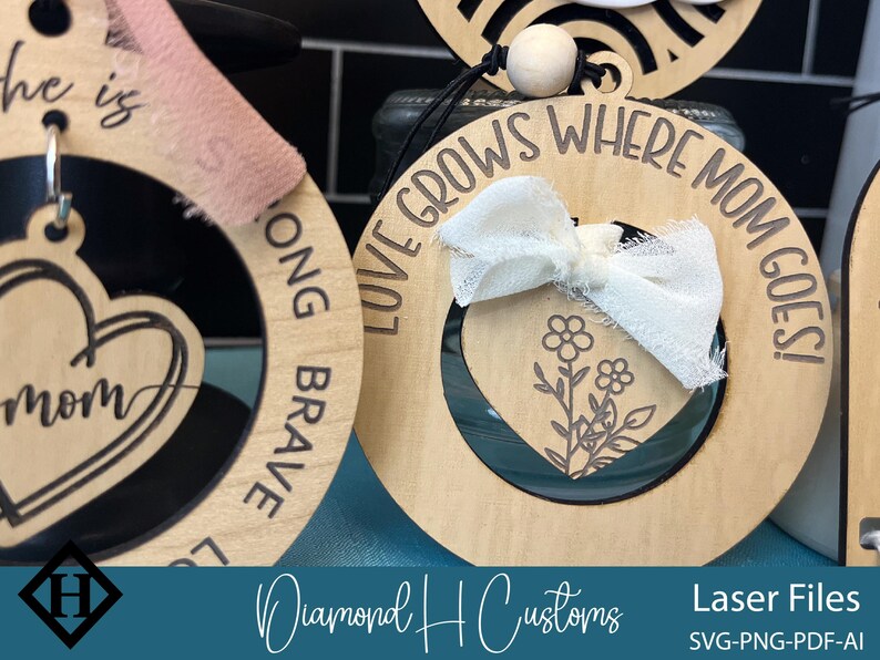 May include: Three wooden ornaments with laser cut designs. The first ornament says "Love Grows Where Mom Goes!" with a white ribbon and a floral design. The second ornament says "Song Brave" with a heart design. The third ornament says "He is Mom" with a heart design.