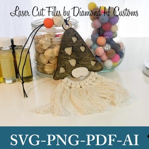 May include: Laser cut wooden gnome ornament with a white tassel beard. The gnome is brown with a white face and has a heart-shaped design with leaves. The ornament is hanging from a black string. The text "Laser Cut Files by Diamond H Customs" is visible at the top of the image.