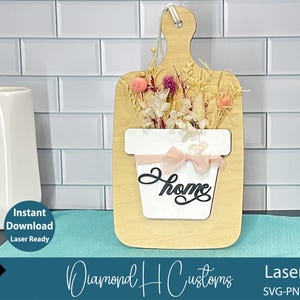 May include: Wooden cutting board-shaped wall decor with a white flower pot design and the word "Home". The pot is filled with dried flowers and accented with a pink ribbon. The sign is laser-ready and available for instant download.