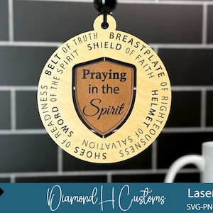 Puede incluir: Un colgante de madera cortado con láser con un diseño circular. El anillo exterior presenta las palabras "Cinturón de la Verdad", "Coraza de la Fe" y otras frases. El centro tiene un escudo con "Praying in the Spirit". El colgante cuelga de un cordón negro.