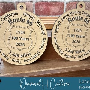 May include: Two wooden Route 66 commemorative ornaments. Each ornament is circular with engraved text including "Route 66", "1926", "100 Years", "2026", and "2,448 Miles". The ornaments are made of light-colored wood.