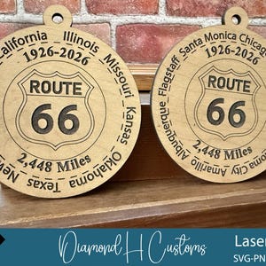 May include: Two wooden Route 66 ornaments. Each ornament features the Route 66 shield logo with the number 66. The text "2,448 Miles" is below the logo. The ornaments are engraved with the names of cities and states along Route 66.