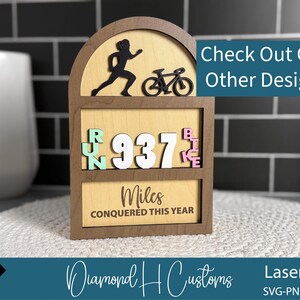 May include: Wooden fitness tracker with a runner and bicycle silhouette. The display shows "RUN 937 BIKE" and "Miles CONQUERED THIS YEAR". The text "Check Out Our Other Designs" is also visible.