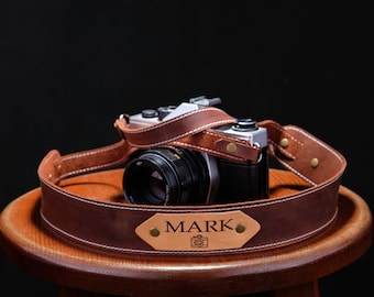 May include: A brown leather camera strap with a personalized nameplate that reads "MARK". The strap is wrapped around a wooden stool and a vintage camera is resting on top of the strap.