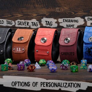 May include: Five different colored leather dice bags with various personalization options. The bags are lined up on a wooden surface with dice scattered in front of them. The options include a gold tag, silver tag, a tag, engraving a name, and engraving an image.