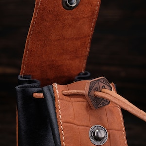 May include: A brown leather dice bag with a black leather bottom. The bag has a drawstring closure with a metal D20 die charm. The bag is open and the inside is visible.