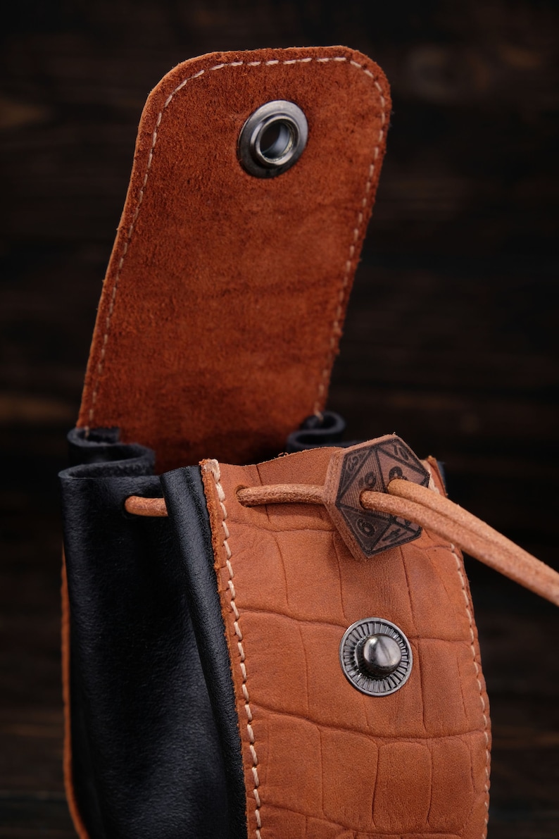 May include: A brown leather dice bag with a black leather bottom. The bag has a drawstring closure with a metal D20 die charm. The bag is open and the inside is visible.