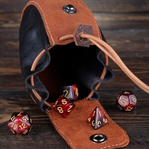 May include: A brown leather dice bag with a black lining and a metal clasp. The bag is open and several red and black dice are spilling out. The dice have gold numbers.