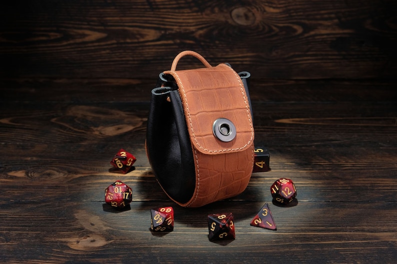 May include: A brown leather dice bag with a black lining and a snap closure. The bag is filled with red and gold dice.