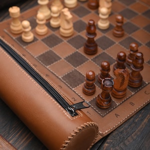 May include: A brown leather chessboard roll with a zipper closure. The chessboard is open and shows a game in progress with white and brown chess pieces.