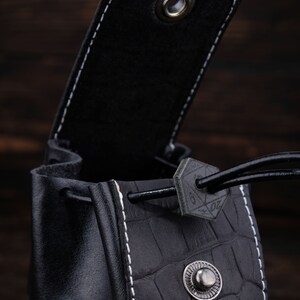 May include: Black leather dice bag with a drawstring closure and a white stitching detail. The bag has a textured crocodile pattern and a metal button.