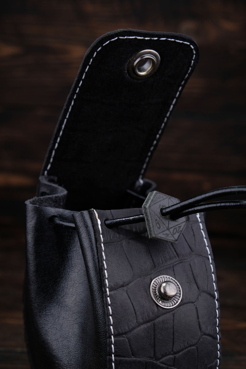 May include: Black leather dice bag with a drawstring closure and a white stitching detail. The bag has a textured crocodile pattern and a metal button.
