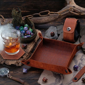 May include: A brown leather dice tray with a crocodile pattern, a brown leather dice pouch with a snap closure, and a brown leather coaster with a glass of whiskey on top. There are also several colorful dice scattered around the tray and coaster.