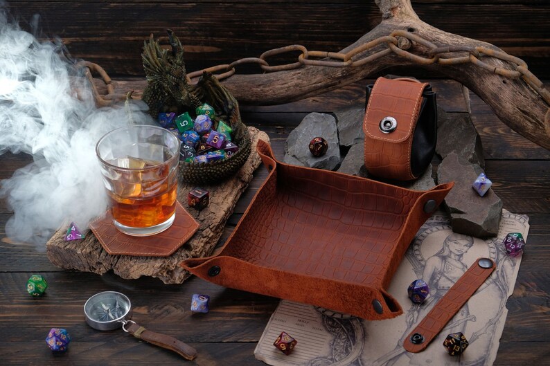 May include: A brown leather dice tray with a crocodile pattern, a brown leather dice pouch with a snap closure, and a brown leather coaster with a glass of whiskey on top. There are also several colorful dice scattered around the tray and coaster.