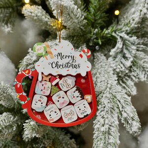 May include: A red Christmas ornament shaped like a mug filled with marshmallows. The ornament has the words "Merry Christmas" and "2025" on it. The marshmallows have faces and names like "Mommy" and "Papa".