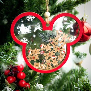 Pode incluir: Um enfeite de Natal vermelho e transparente em forma de orelhas do Mickey Mouse. O enfeite apresenta uma borda vermelha, flocos de neve, o trenó do Papai Noel, renas e bonecos de gengibre. O texto "The Smith Family" é visível.