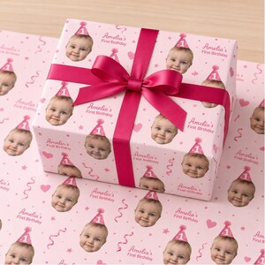 May include: A rectangular gift box wrapped in light pink paper featuring a repeating pattern of a baby wearing a pink party hat with the number one, pink hearts, streamers, and the text Amelia's First Birthday, tied with a dark pink satin ribbon.
