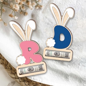 May include: Two wooden bunny-shaped money holders with slots for bills. The bunny on the left is pink with the letter "R" and the bunny on the right is blue with the letter "D".