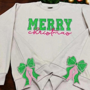 Pode incluir: Camisola branca com o texto "Merry Christmas" em glitter verde e laços de glitter rosa nas mangas.